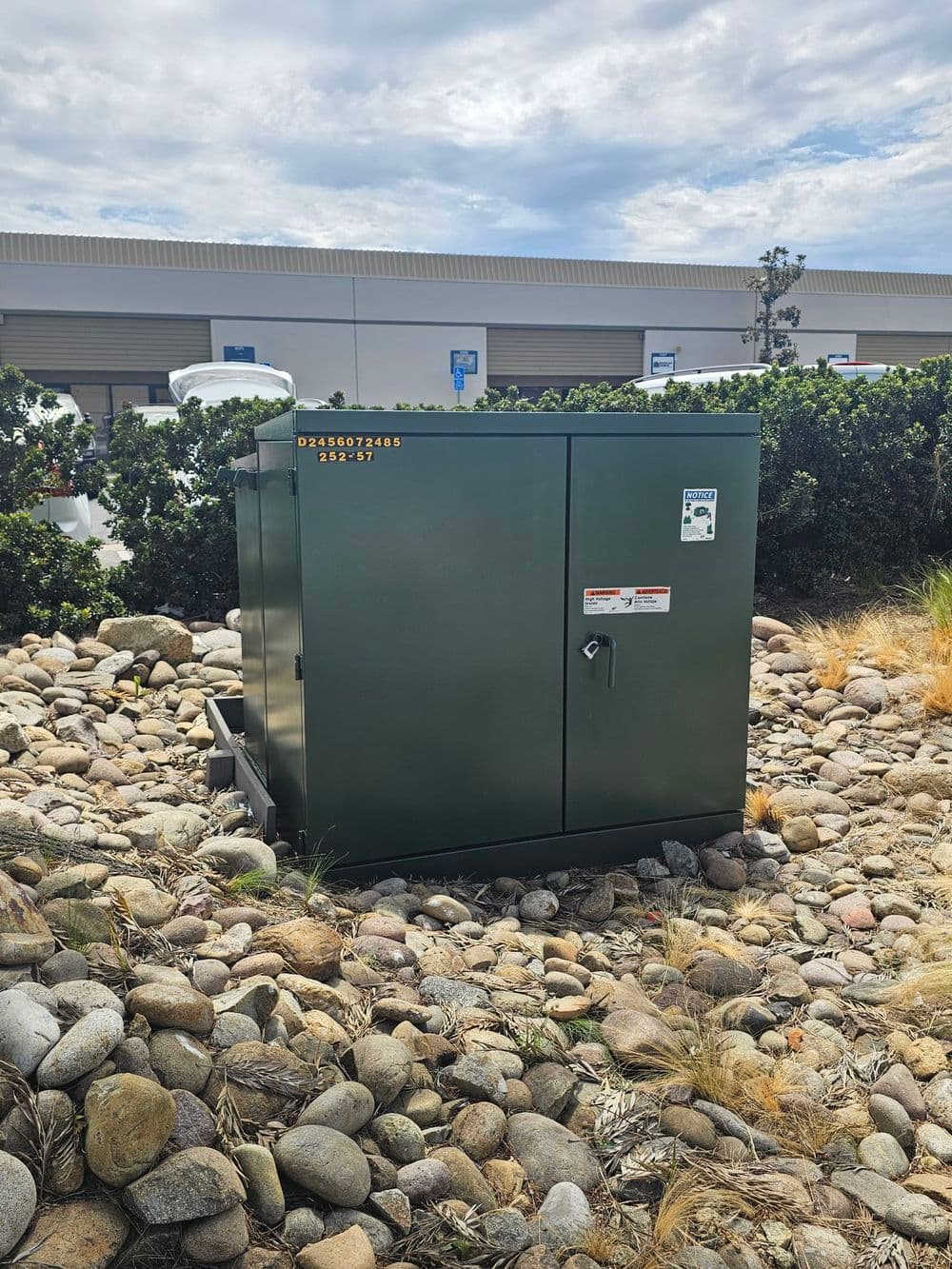 Green utility cabinet surrounded by rocks and bushes under a cloudy sky.