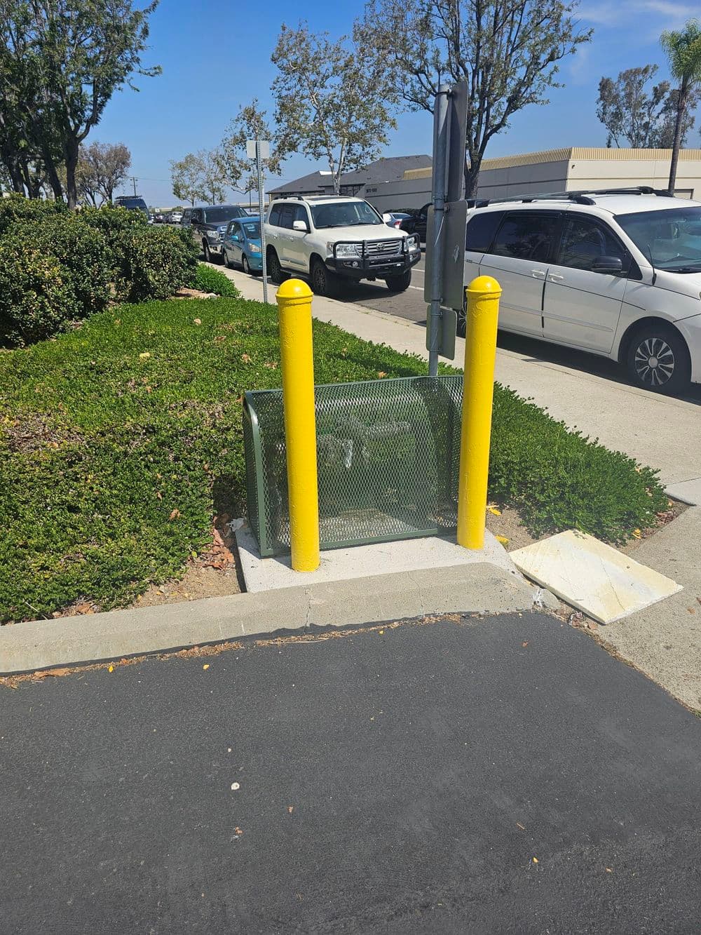 Yellow bollards protect a mesh trash enclosure near parked cars and greenery.